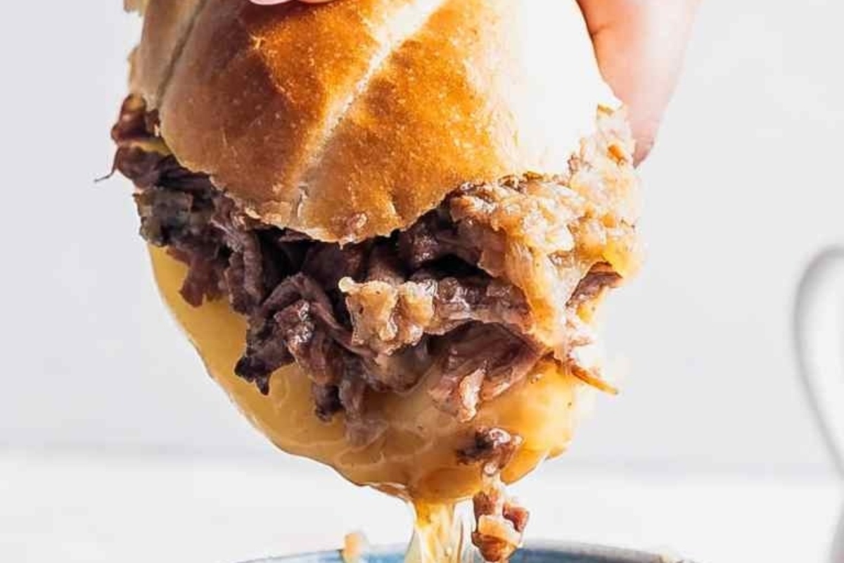 A person is serving a sandwich on a bun and dipping it into a bowl of sauce.