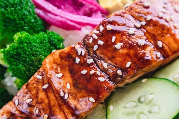 A budget-friendly takeout meal featuring a plate of salmon, broccoli, and cucumbers sprinkled with sesame seeds.