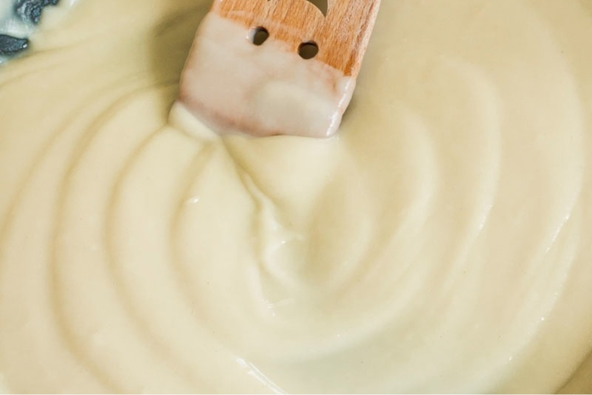 A wooden spoon is being used to stir a bowl of white sauce in a recipe.