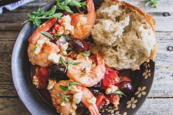 A bowl of shrimp with tomatoes, olives and bread.