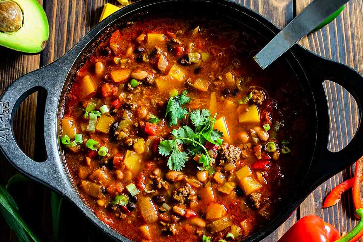 A bowl of chili with vegetables and avocado on a wooden table.