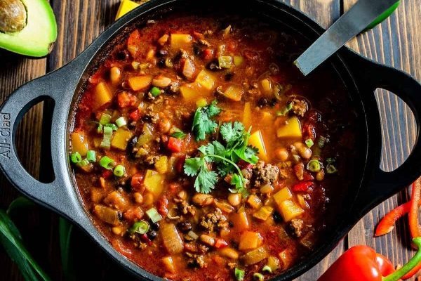 A bowl of chili with vegetables and avocado on a wooden table.