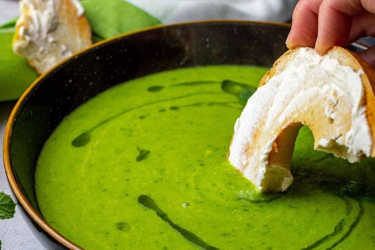 A person is dipping a bread into a bowl of green soup made with frozen peas.