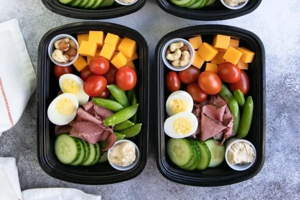 Four black plastic containers filled with ham, eggs, tomatoes, and cucumbers prepared for lunchtime meal prep.