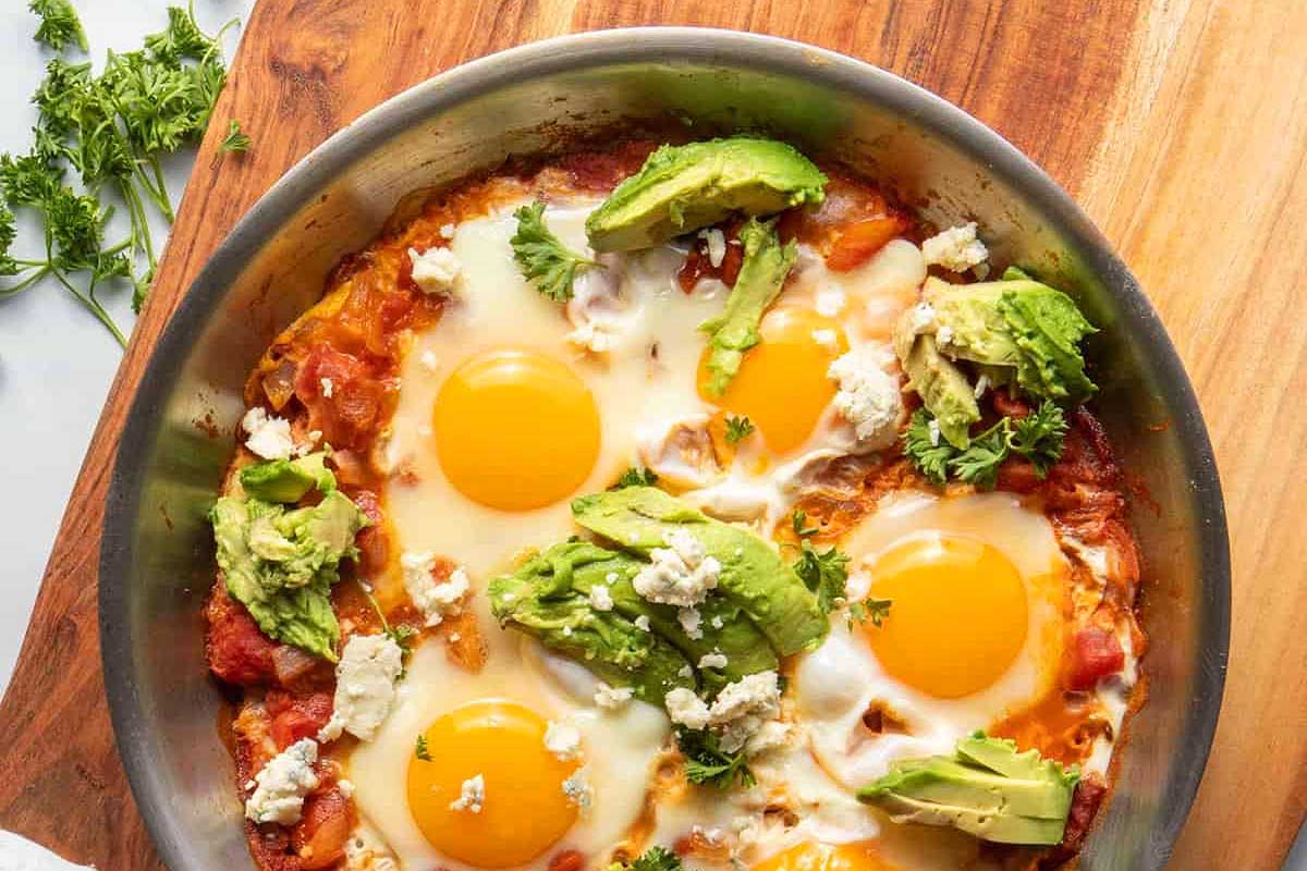 A skillet with eggs and avocado on a wooden cutting board.