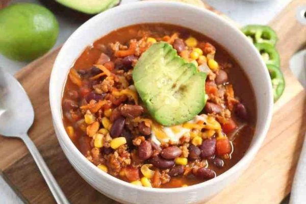 A bowl of chili with corn, beans and avocado on a wooden cutting board.