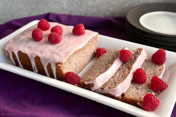 A slice of raspberry bread with icing and raspberries on a white plate.