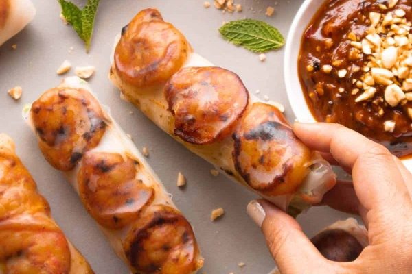 A budget-friendly person is holding a plate of grilled banana spring rolls with dipping sauce.