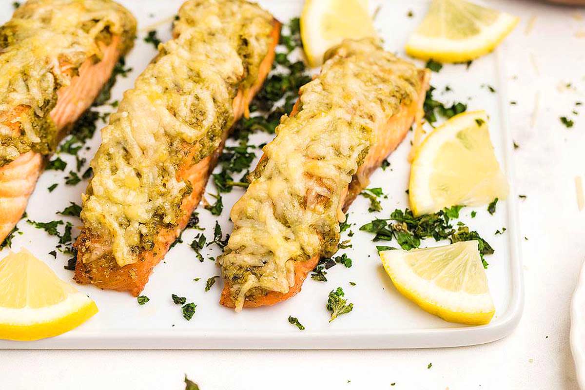 Three salmon fillets on a white plate with lemon wedges, drizzled with a flavorful pesto sauce. This is one of the best pesto recipes.
