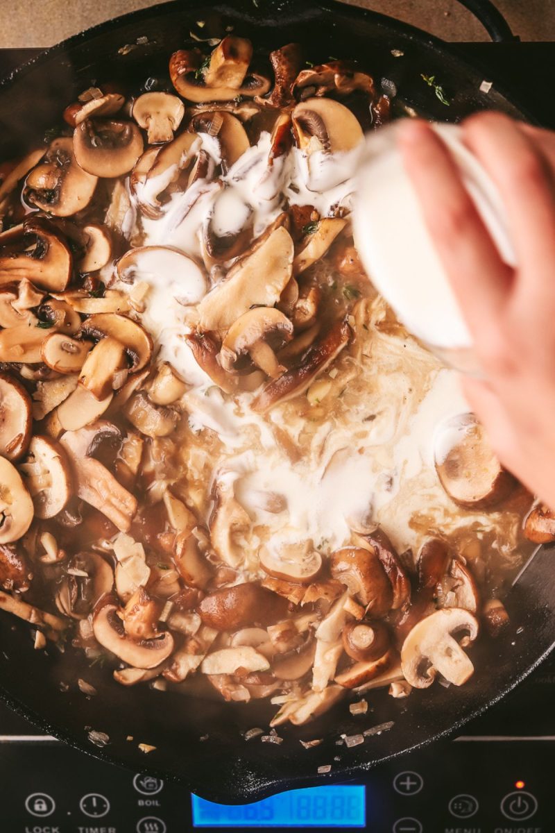 A person pouring champagne into a frying pan with mushrooms.