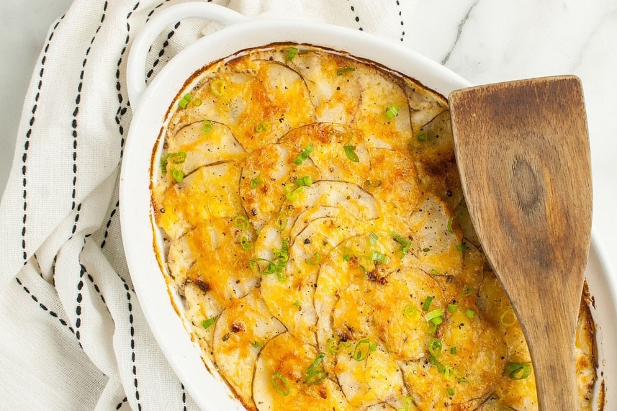 A cheesy potato casserole served as a delicious side dish, presented in a white dish and accompanied by a wooden spoon.