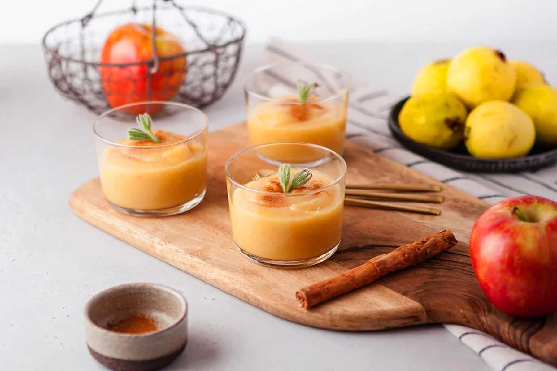 A glass of orange juice with cinnamon sticks and apples on a cutting board.