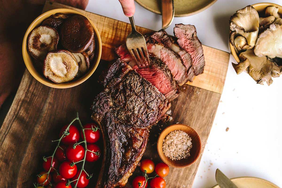 A person cutting a steak on a cutting board with tomatoes and mushrooms.