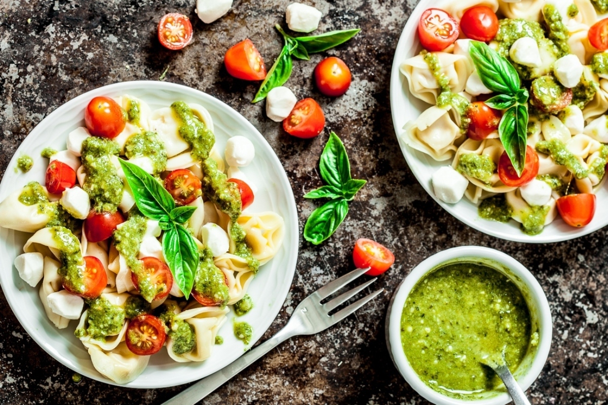 Two plates of pasta with pesto sauce, served with sides of tomatoes.