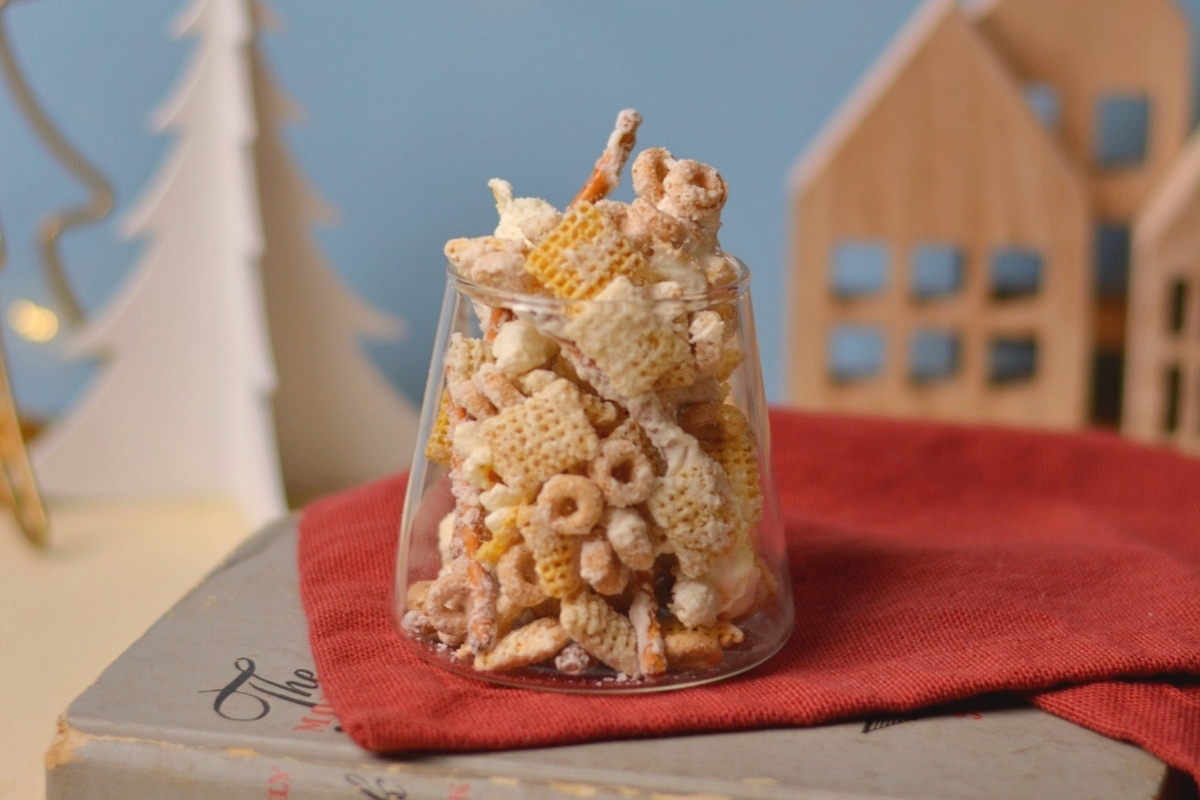 A tantalizing combination of crispy Chex mix, coated in creamy white chocolate, presented elegantly in a glass nestled upon a book.