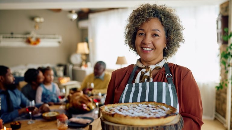 Happy mature woman holding baked Thanksgiving pie and looking at camera. Her family is in the background.