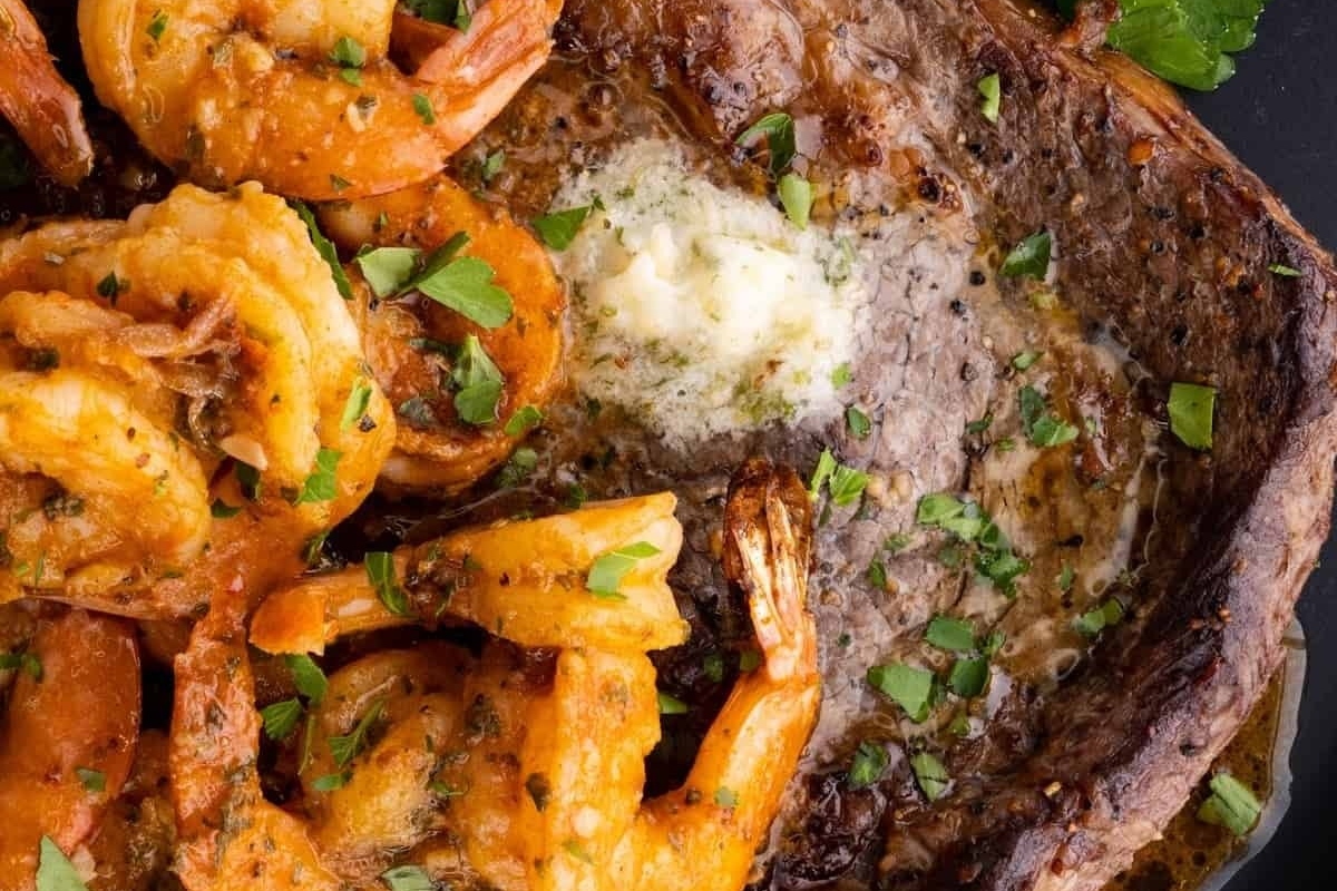 A surf and turf platter consisting of a flavorful steak, succulent shrimp, and aromatic herbs served on an elegant black plate.