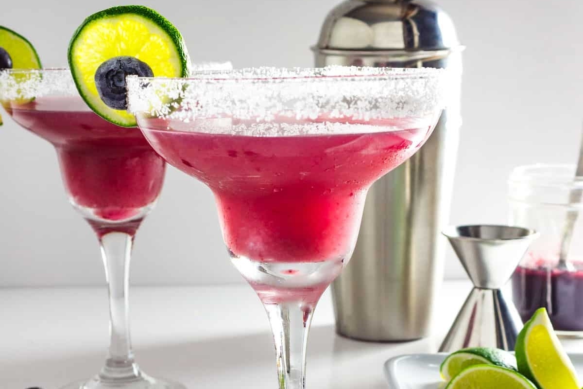 Two Valentine's Day cocktails in margarita glasses with blueberries and limes.