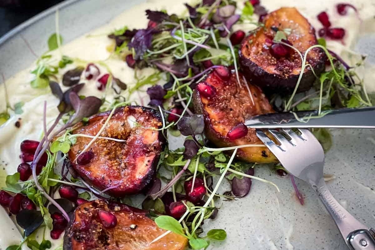 A plate of figs and pomegranate with a fork, accompanied by goat cheese appetizers.