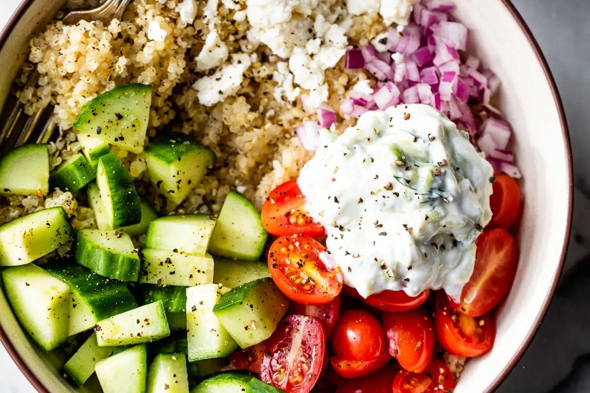 A grain bowl of quinoa salad with cucumbers, tomatoes, and sour cream.