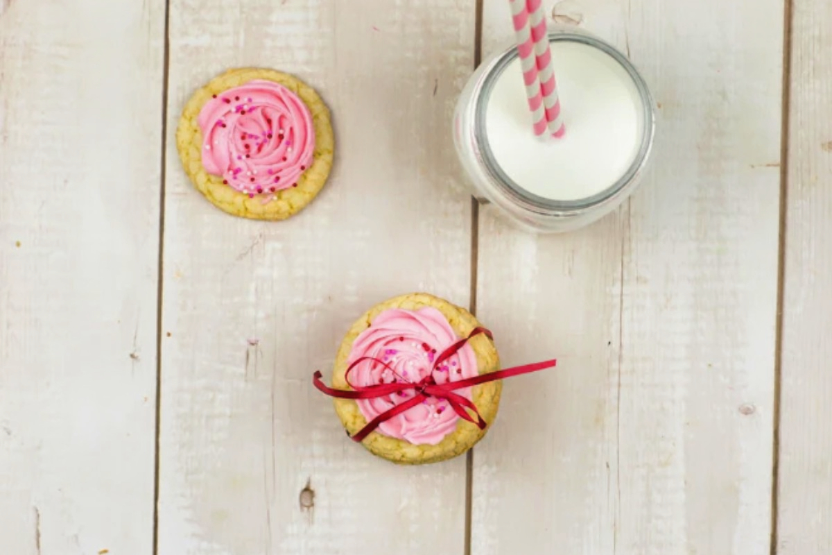 Pink frosted cookies on a wooden table next to a glass of milk, perfect for Valentine's Day.