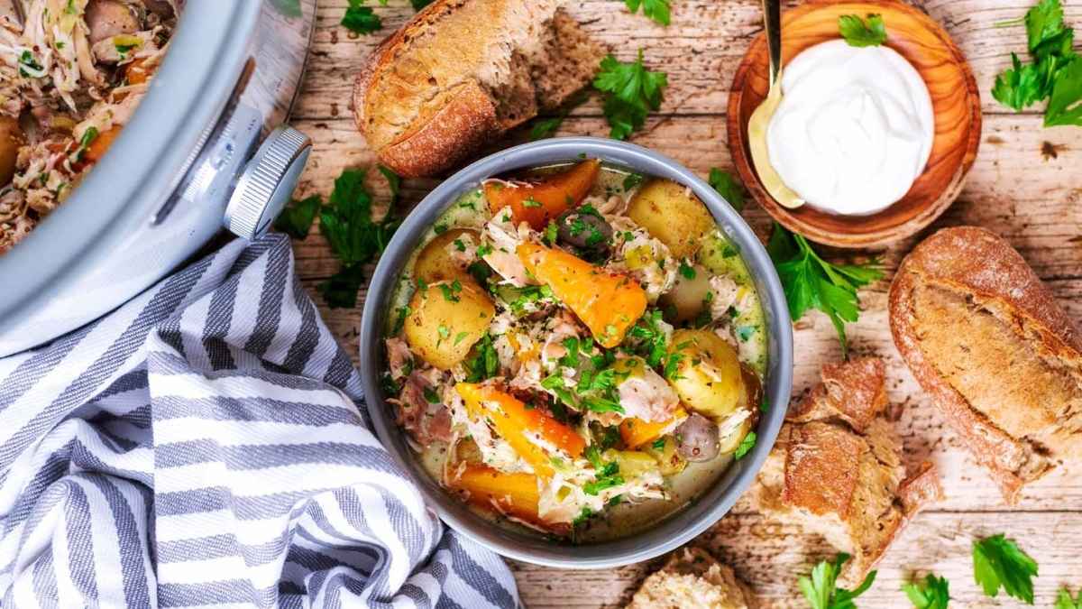 A slow cooker dinner of chicken stew with potatoes and bread on a wooden table.