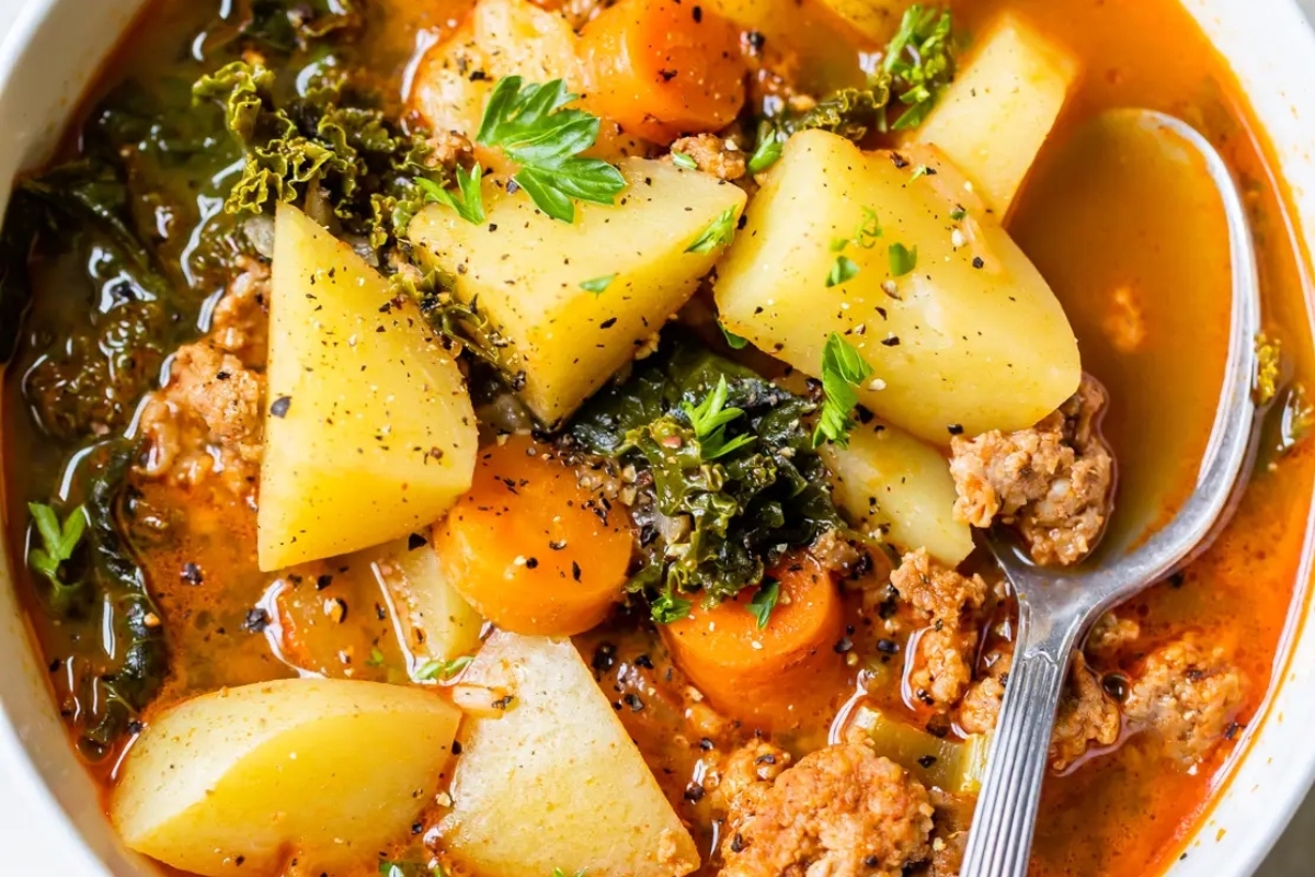 A bowl of potato soup with meat, potatoes and kale.