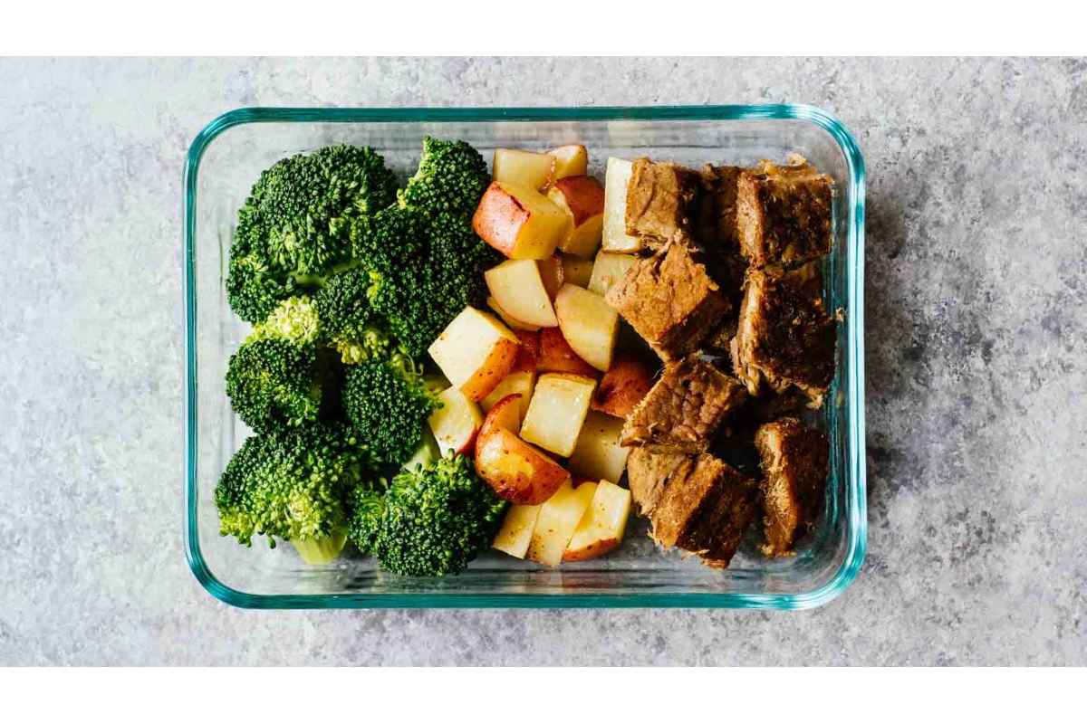 A healthy glass container filled with high protein meat, nutritious broccoli, and fresh apples.
