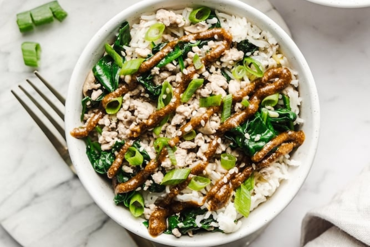 A white bowl with rice, greens, and pork in it.