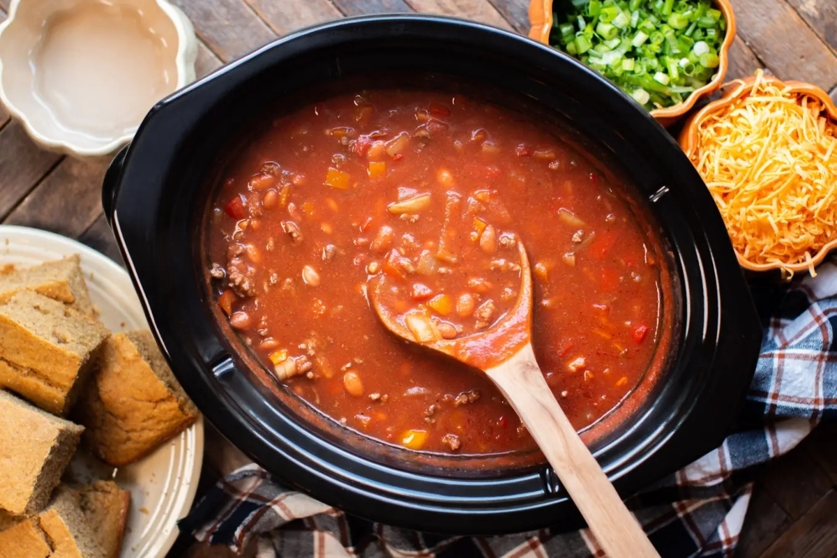 A slow cooker full of chili and bread on a wooden table.