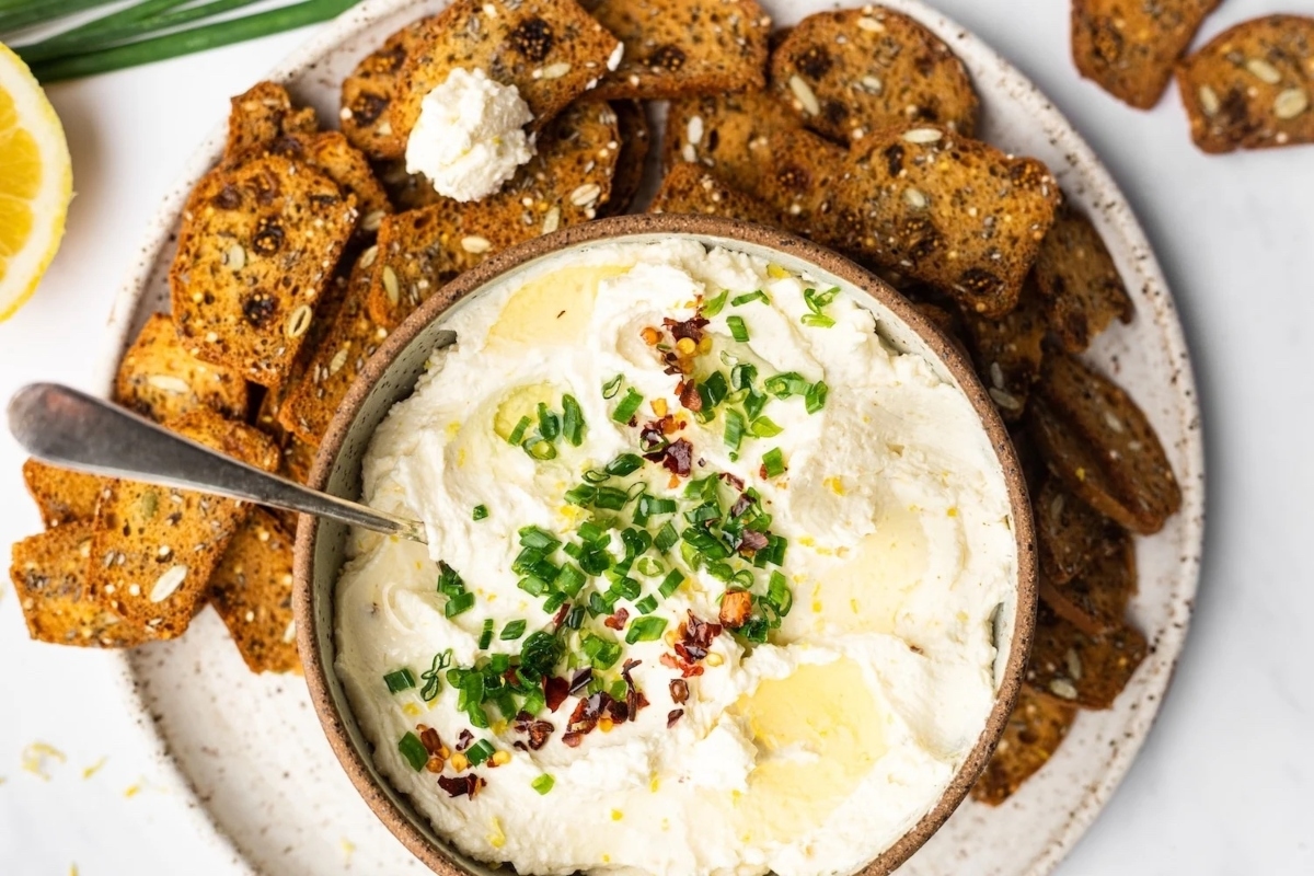 A creamy dip made with cream cheese, served in a bowl alongside crispy crackers on a white plate.