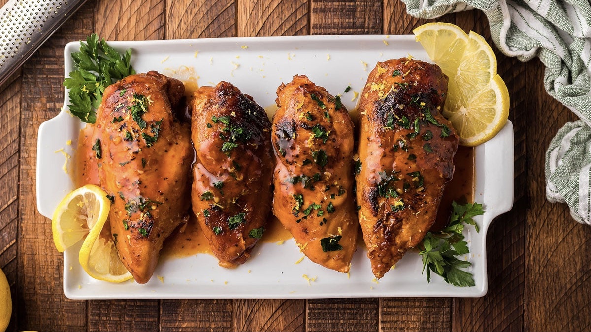 Four chicken breasts on a white plate with lemon wedges.