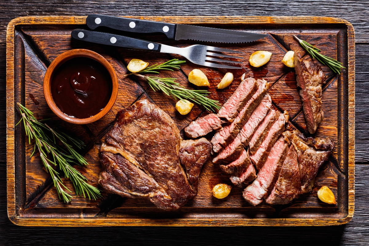 Fried juicy chuck eye beef steaks, a whole and one cut in slices with fried garlic cloves and rosemary on a rustic rude wood board with barbecue sauce on a wooden table with cutlery.