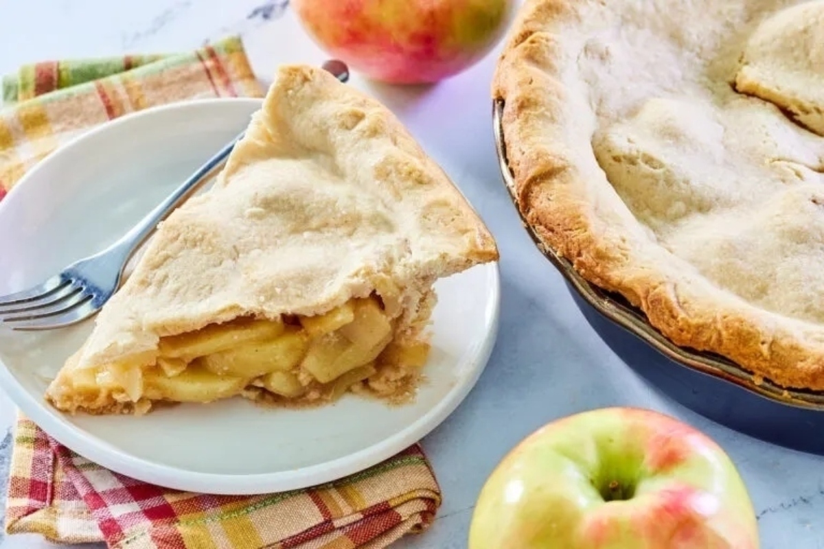 A slice of apple pie on a plate next to apples.