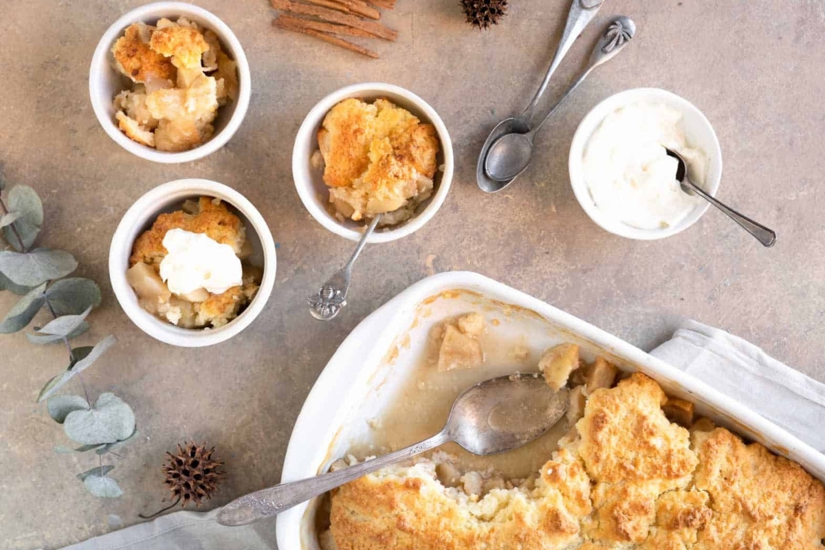 Apple cobbler in a white dish with whipped cream and cinnamon sticks.
