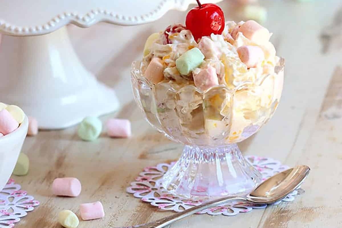 A bowl of fruit salad topped with a cherry, surrounded by marshmallows, with a spoon on the side.