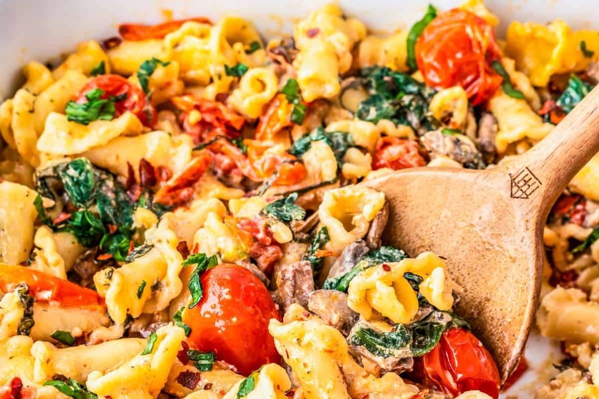 Cooked pasta with tomatoes, spinach, and sauce in a dish with a wooden spoon.
