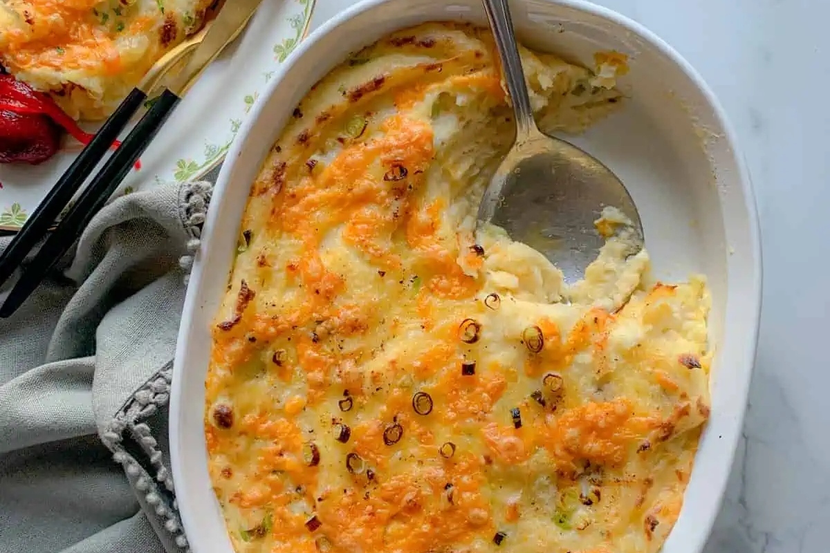 A casserole dish filled with baked cheesy mashed potatoes, partially scooped, with a spoon inside.