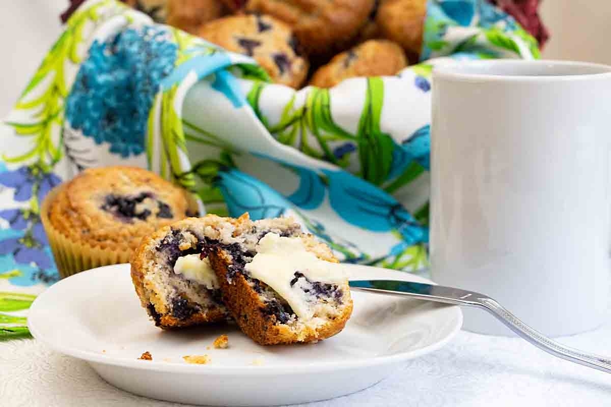 A plate with a halved blueberry muffin and a fork, next to a white mug, with more muffins and a colorful napkin in the background.