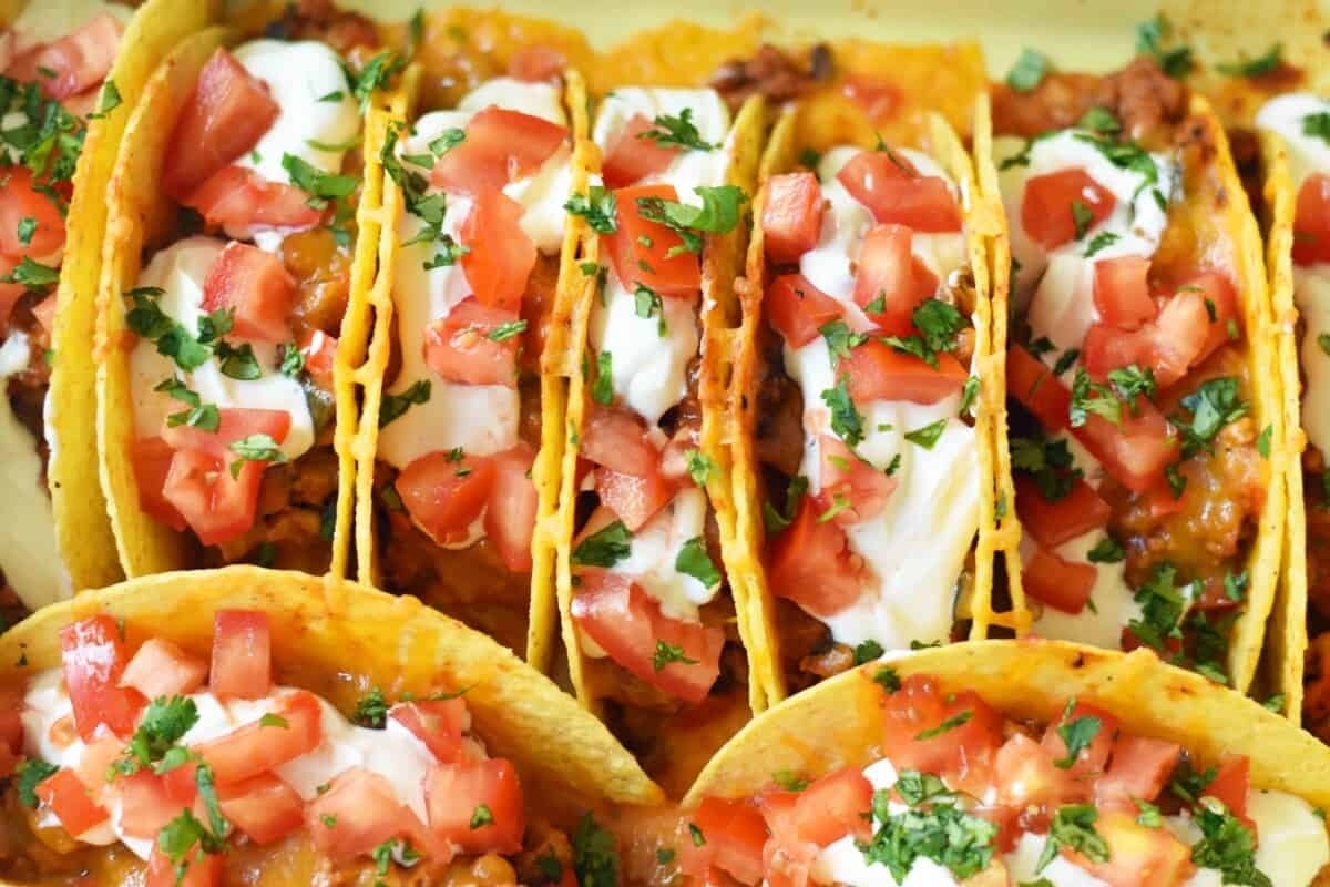 A row of hard-shell tacos filled with ground meat, topped with sour cream, diced tomatoes, and sprinkled with chopped parsley.