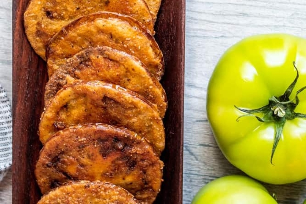 Fried plantain slices served on a wooden platter next to green tomatoes.
