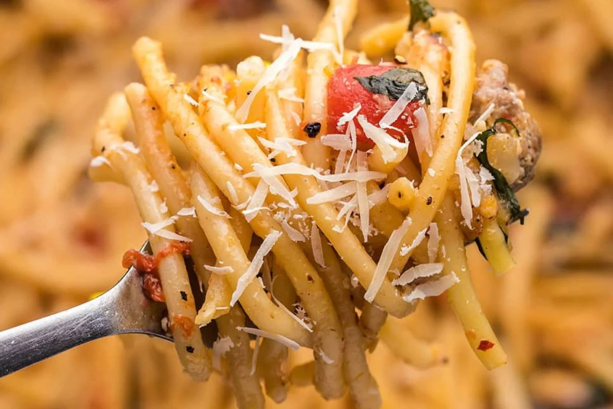 Close-up of a forkful of pasta topped with grated cheese, tomato pieces, and herbs, against a blurred background of more pasta. Perfect for recipes featuring noodles.