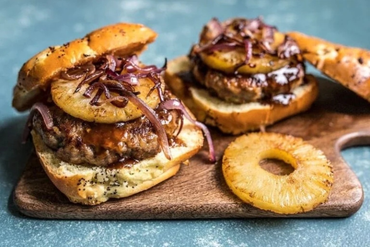 Two burgers with pineapples and onions on a wooden cutting board.