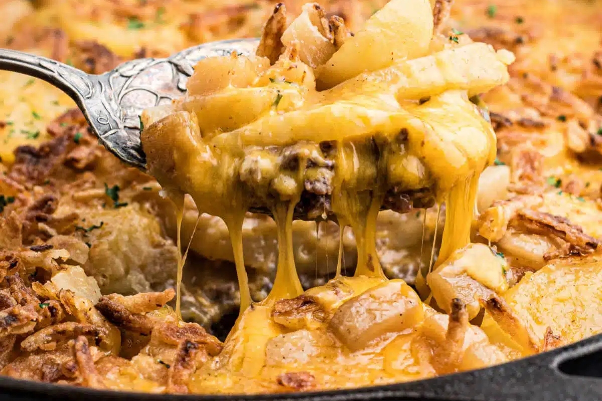 A close-up of a cheesy potato dish being served with a spoon, showing melted cheese stretching from the pan.