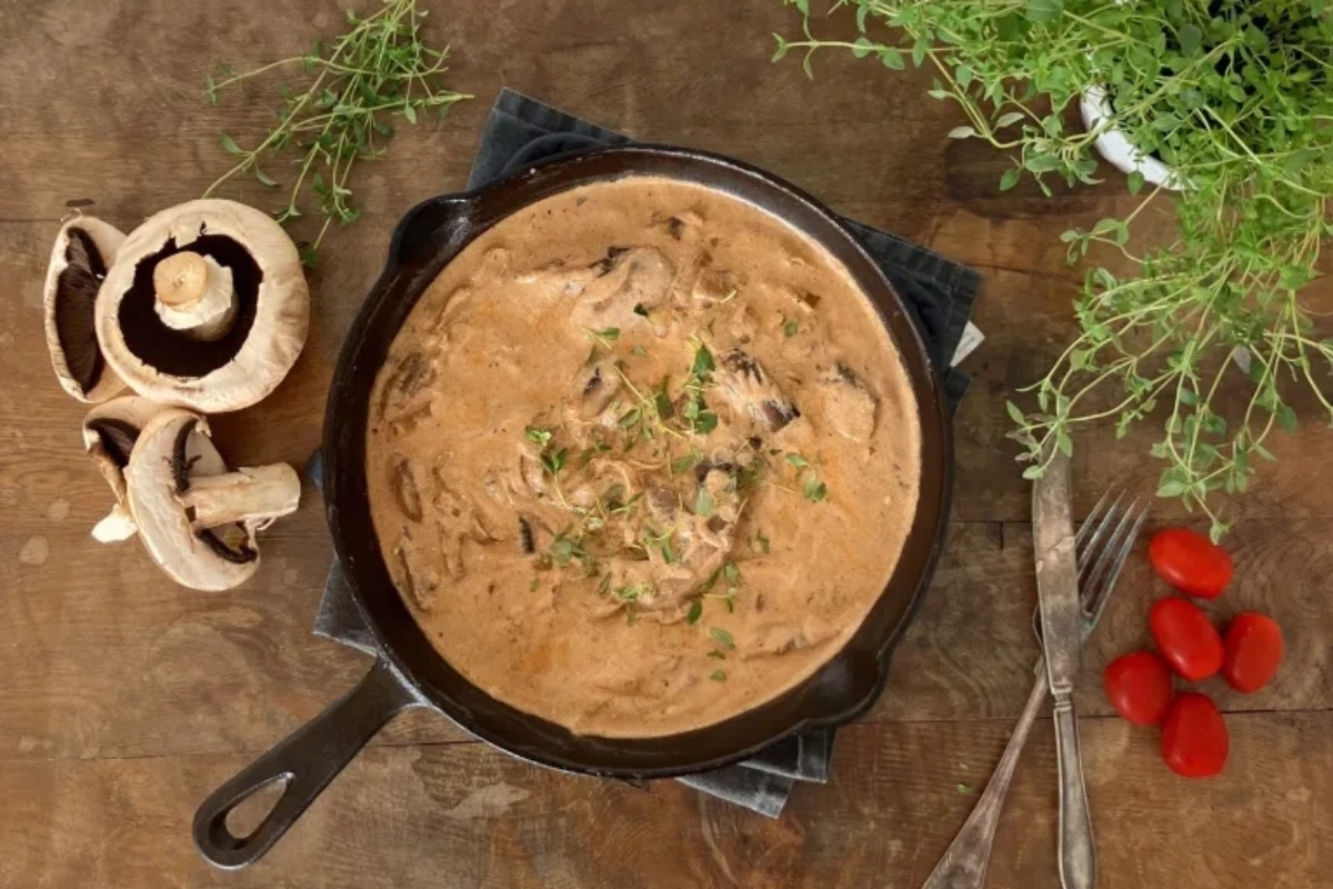 Cast-iron skillet with creamy mushroom sauce, garnished with herbs, surrounded by fresh ingredients on a wooden table.