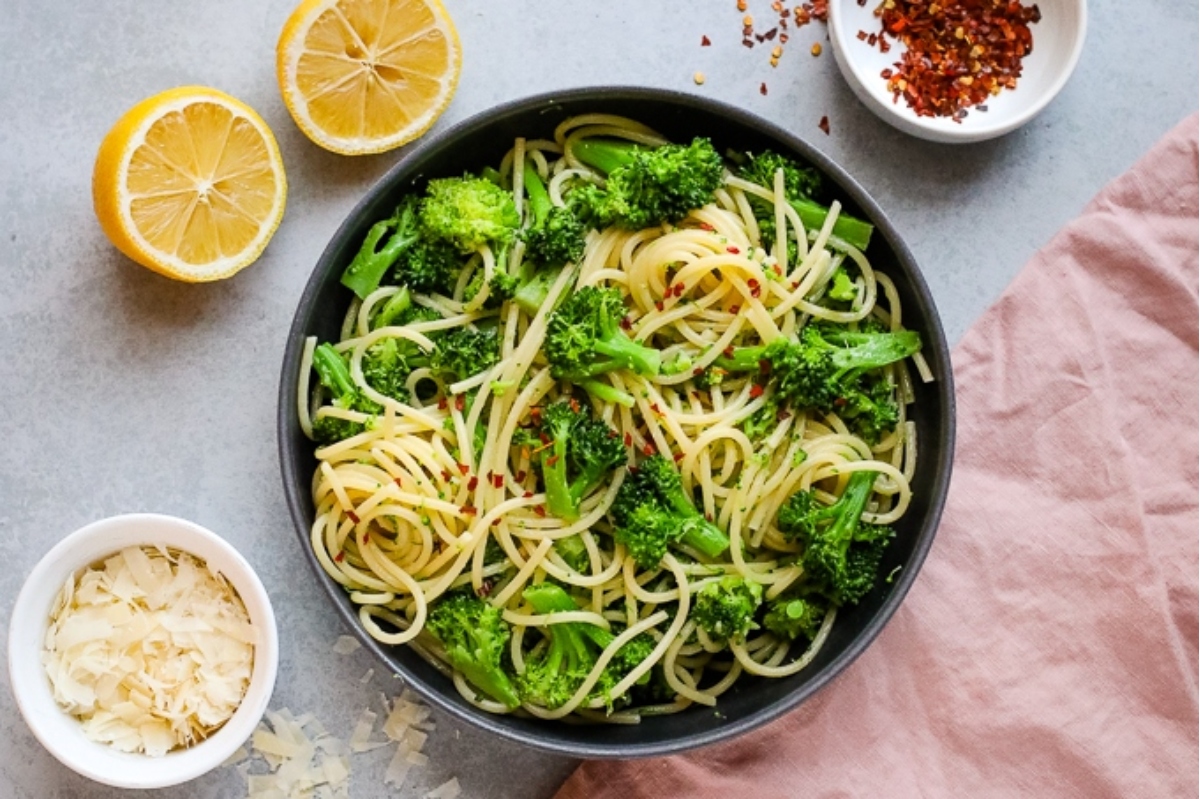 A bowl of pasta with broccoli, garnished with lemon wedges and red chili flakes, alongside a small bowl of grated cheese.