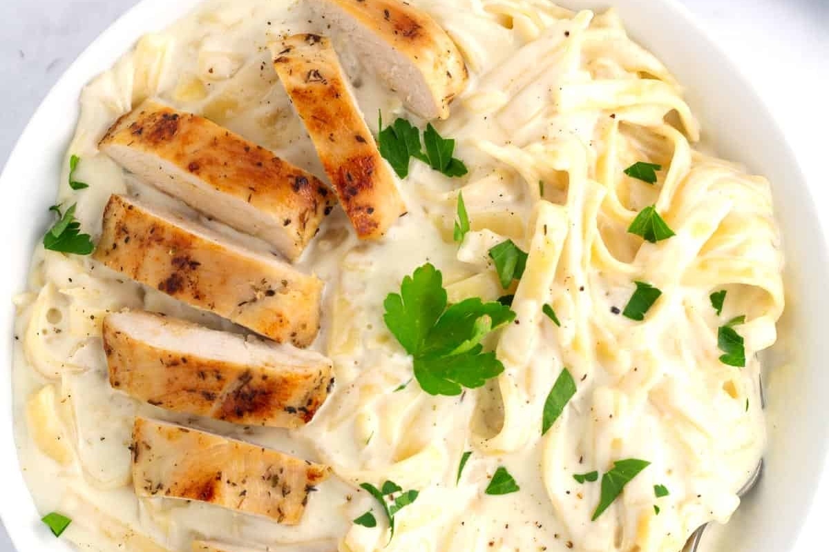 A plate of creamy fettuccine alfredo topped with grilled chicken strips and garnished with parsley.