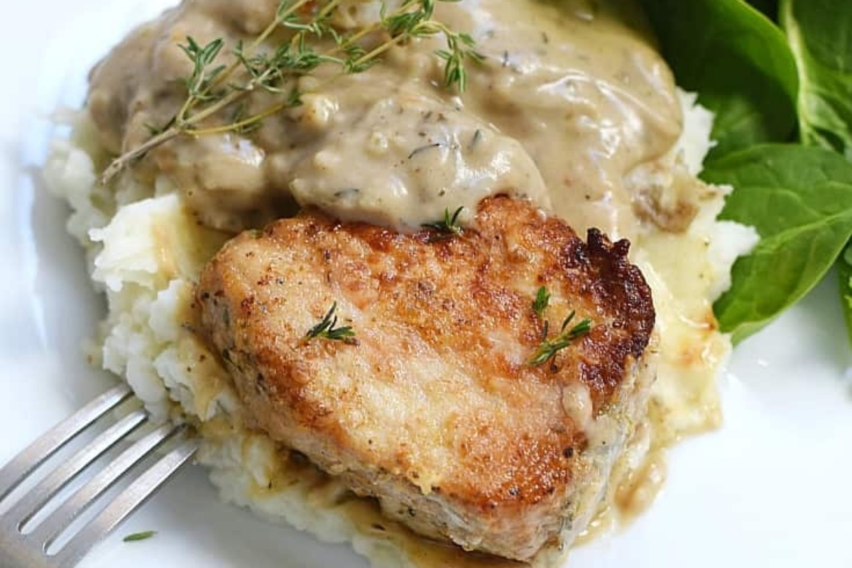 Seared pork chop served over mashed potatoes with creamy mushroom sauce and a garnish of fresh herbs.