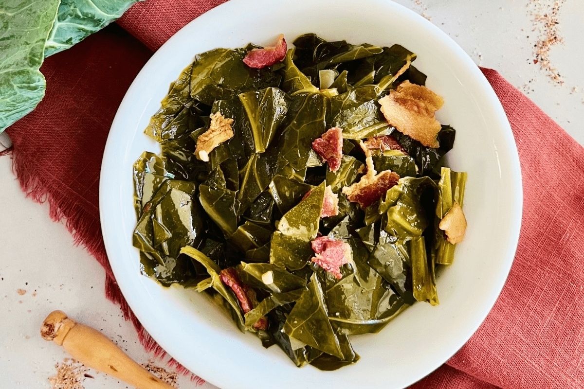 A bowl of cooked collard greens with bits of bacon and garlic, served on a table with a napkin and wooden spoon nearby.