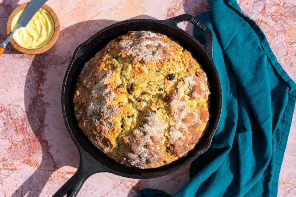 A freshly baked loaf of bread in a cast-iron skillet, accompanied by a small bowl of butter, on a marbled pink surface with a teal cloth.